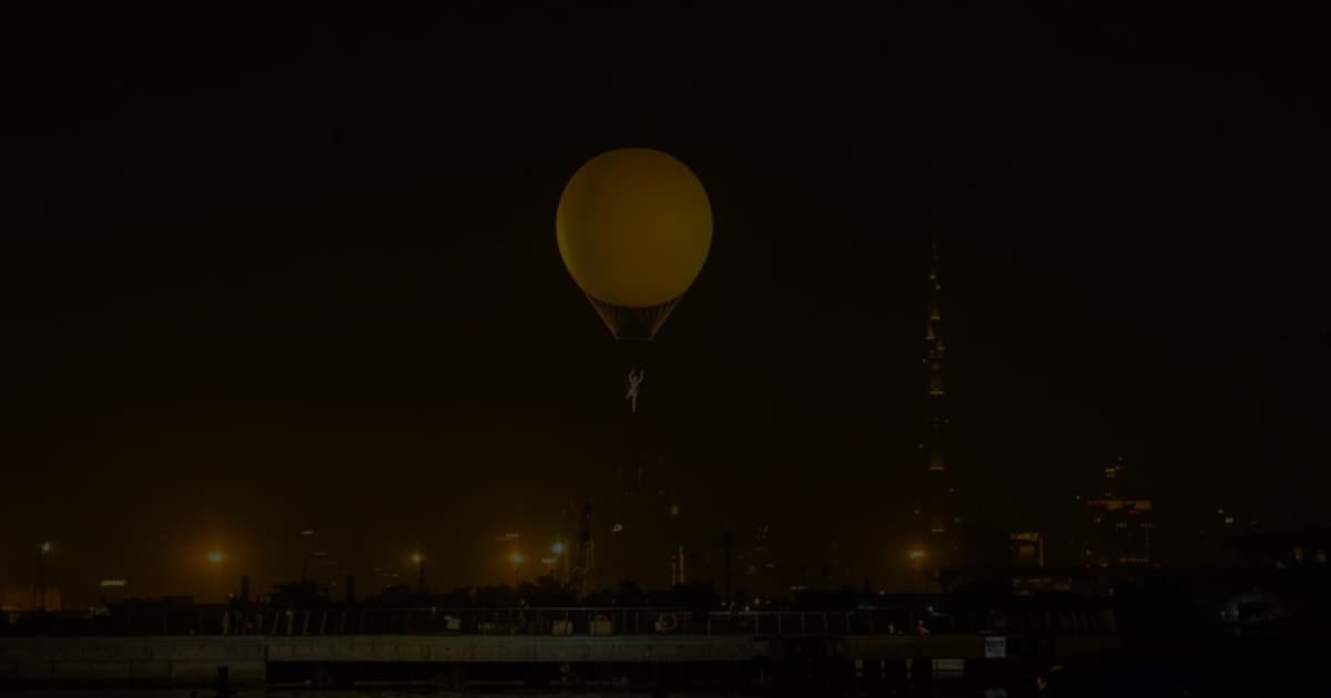 Distant view of the illuminated helium balloon and aerialist over Dubai Creek at night with the city skyline behind